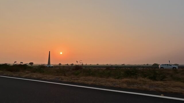 A scenic wide driving shot along an empty highway asphalt road during a beautiful golden hour sunset. The sun glows through a hazy orange sky over rural fields.
