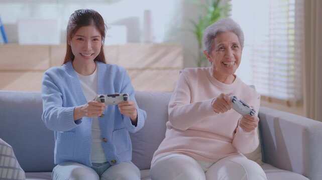 Happy senior mother and adult daughter playing video games together on a sofa at home.  intergenerational fun, gaming, living room leisure, and modern retirement lifestyle.  family bonding Concept