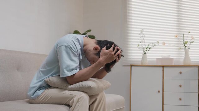 An Asian man, looking stressed about household finances, slumps down in his living room, showing signs of worry.