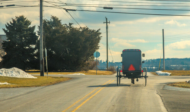 A horse-drawn carriage moves down a country road. The area is quiet with patches of snow on the ground. Trees line the sides of the road. The sky is partly cloudy.