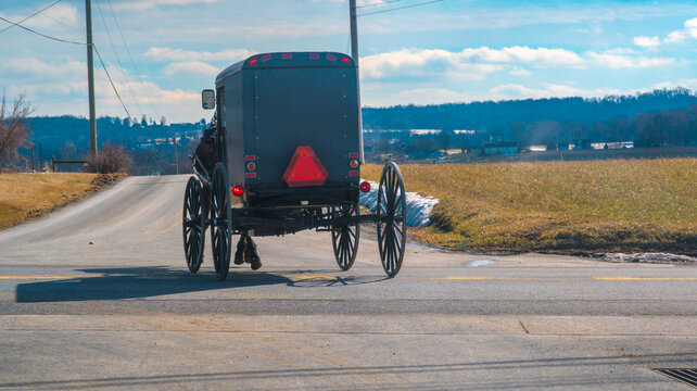 A horse-drawn buggy moves along a country road lined with grass and fields. The sky is clear with some clouds. The scene shows rural life in a quiet area.