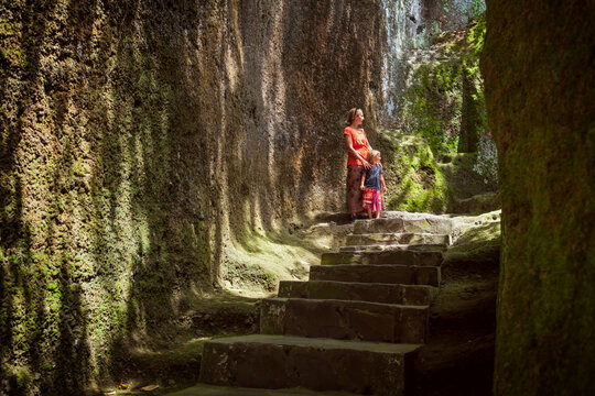Mother and children in sarongs walk near ancient rock temple ruins in Gunung Kawi, Bali &mdash; exploring spiritual heritage and enjoying a cultural family adventure in lush tropical nature.