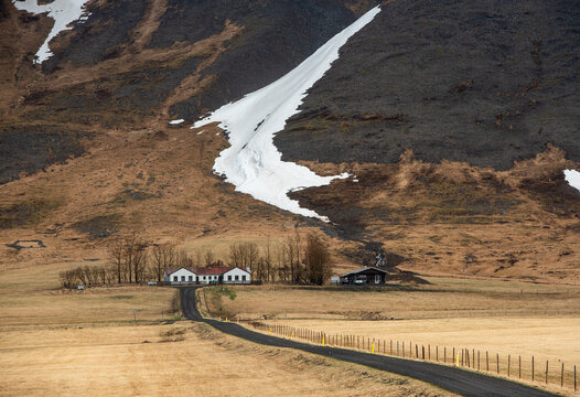Winding country road leading to a remote farmhouse under a massive mountain with melting snow in Iceland.