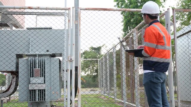 Professional male electrical engineer uses a digital tablet while safely inspecting a high-voltage power transformer outside a modern commercial energy facility managing grid infrastructure.