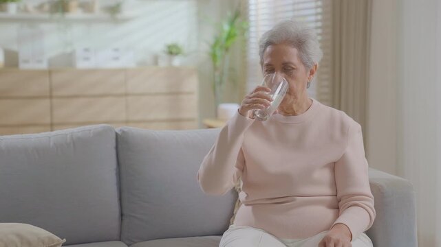 Olde retired or senior elderly asian woman drinking a glass of water on couch at home. Drink enough water. relaxing sitting on sofa at day time. healthy hydration, lifestyle independent living