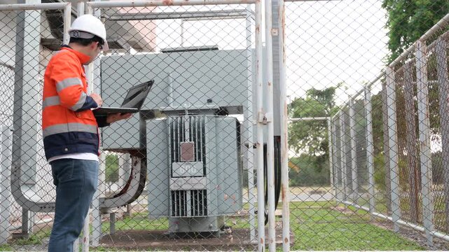 Professional male electrical engineer uses a digital tablet while safely inspecting a high-voltage power transformer outside a modern commercial energy facility managing grid infrastructure.