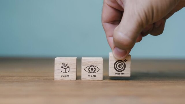 Close-up of a hand placing wooden blocks with Values, Vision, and Mission text and icons, representing corporate strategic planning, business identity, core belief, and target goal concept.