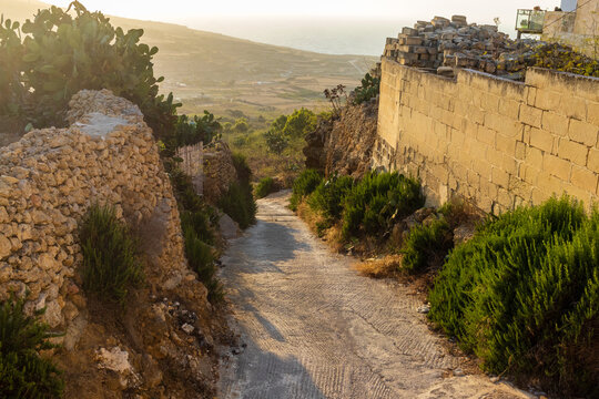
Sunlit Stone Path Through Rural Mediterranean Village Between Dry Stone Walls Overlooking Coast