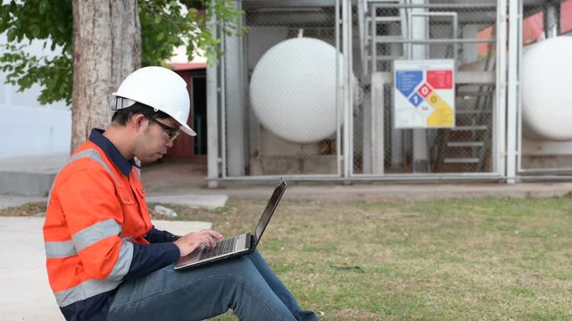 Professional male industrial engineer uses a digital tablet while standing outside inspecting large white chemical storage tanks at a modern energy manufacturing facility during the day.