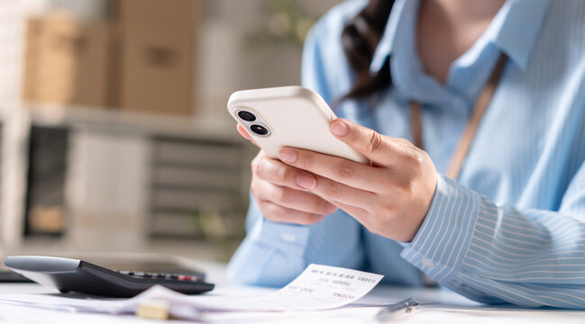 Close-up of female employee hand using smartphone with financial calculator and invoice receipt in office. Professional business woman checking mobile payment and online banking data.