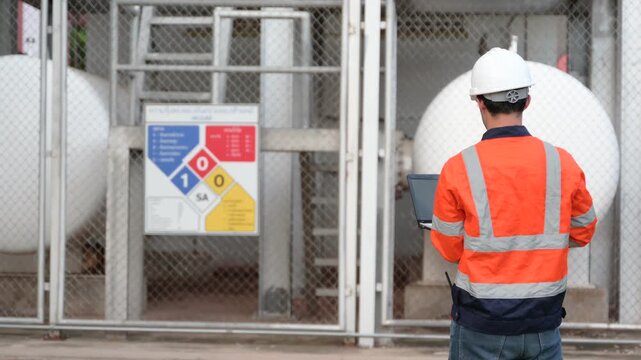 Professional male industrial engineer uses a digital tablet while standing outside inspecting large white chemical storage tanks at a modern energy manufacturing facility during the day.