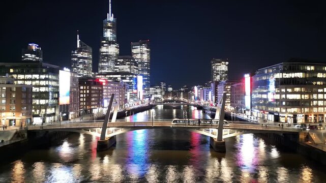 Modern city skyline with a tram crossing a bridge over a river at night.