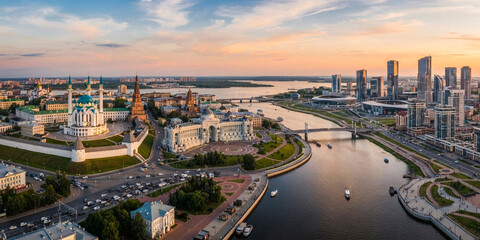 Fototapeta premium A wide-angle aerial view of a city center at sunset, showcasing a prominent mosque with blue domes next to a river and modern skyscrapers