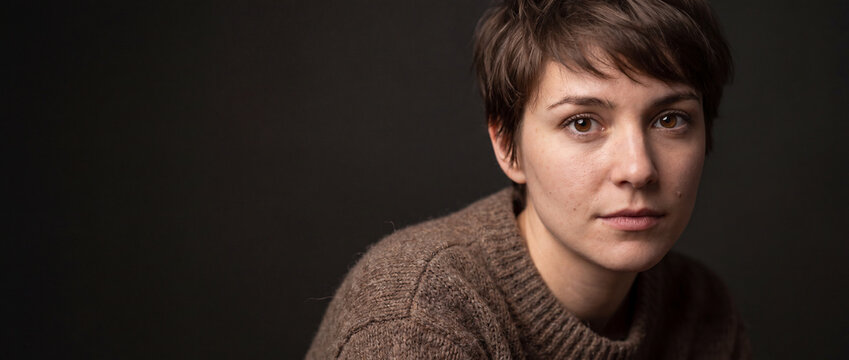 Portrait of a young woman with short hair wearing a brown knitted sweater, captured in close-up against a dark studio background, ideal for lifestyle and mental health concepts