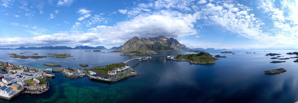 A breathtaking wide aerial panorama captures the islands of Henningsv&aelig;r in Lofoten, Norway, linked by bridges over turquoise waters under dramatic mountain peaks and a cloud-streaked blue sky.