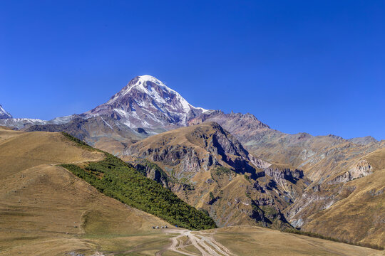 Majestic Mount Kazbek rises behind Gergeti Trinity Church in Georgia. High angle view in bright autumn afternoon light