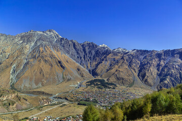 Fototapeta premium High angle view of Stepantsminda town in the Caucasus mountains, Georgia. Scenic panorama from a hill in sunny daylight