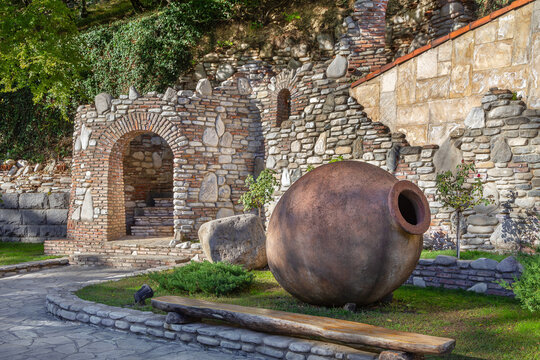 Big clay qvevri vessels lie in a green garden by a stone wall, Georgia. Entrance to a traditional winery in daylight