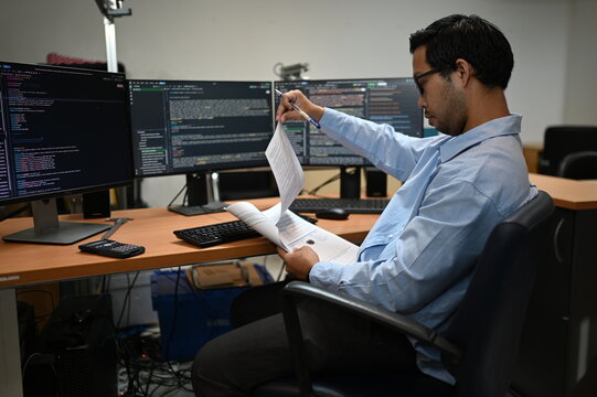 Focused Asian male software developer types on a keyboard analyzing complex programming code on multiple computer monitors while working at a desk inside a modern corporate IT business office.