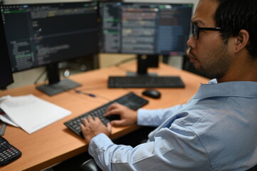Fototapeta premium Focused Asian male software developer types on a keyboard analyzing complex programming code on multiple computer monitors while working at a desk inside a modern corporate IT business office.