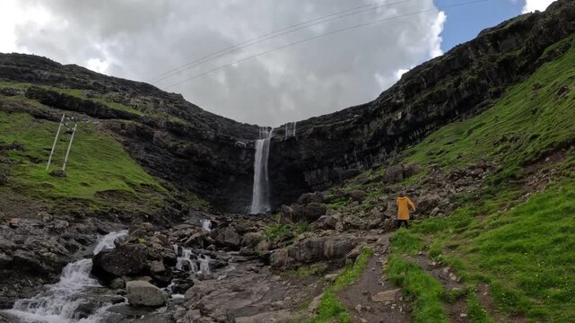 Tourist in yellow raincoat walking towards Fossa waterfall, Faroe Islands