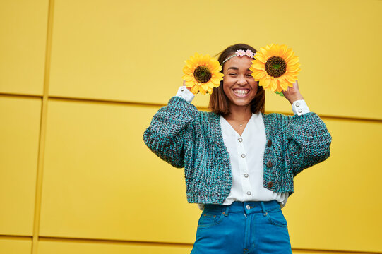 Cheerful young woman is posing with sunflowers in front of a yellow wall
