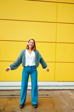 Carefree young woman listening to music and dancing with pink headphones in front of yellow wall