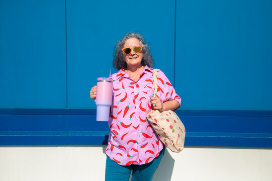 Senior woman with colorful clothes and reusable bottle is posing in front of a blue wall
