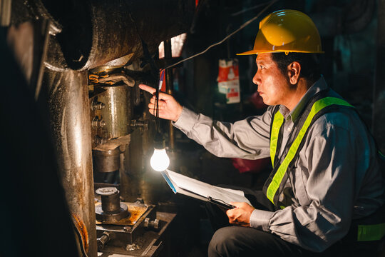 Engineer helmet safety vest inspection machine factory worker clipboard industrial maintenance male technician examining heavy equipment with lamp during night shift in dim manufacturing workshop