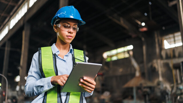 Engineer worker tablet helmet vest factory industry woman safety inspection, confident female industrial worker using digital tablet in factory while wearing blue hard hat and green safety vest