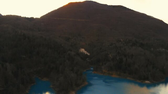 Horizontal 4K 24fps aerial drone video starting high on dark winter mountains and slowly panning left to reveal the beautiful turquoise water of a lake, with a bright sunlit peak in the backgroun