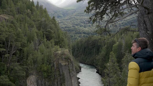 Man in a yellow and black jacket standing by a tree, observing a river flowing through a deep forest canyon.
