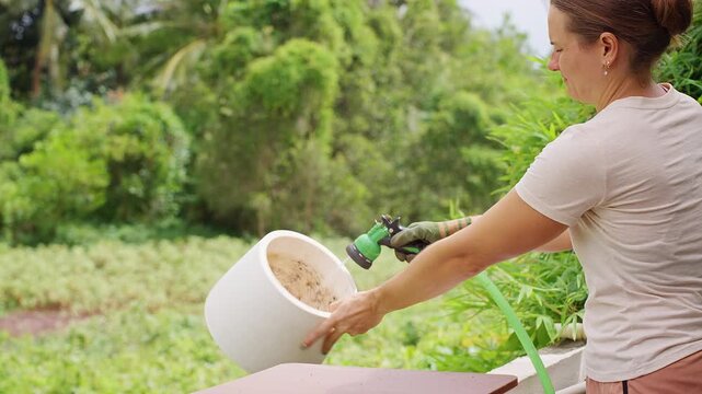 Woman Rinses an Empty White Plant Pot with a Garden Hose, Cleaning the Container Outdoors before Reuse for Sustainable Gardening
