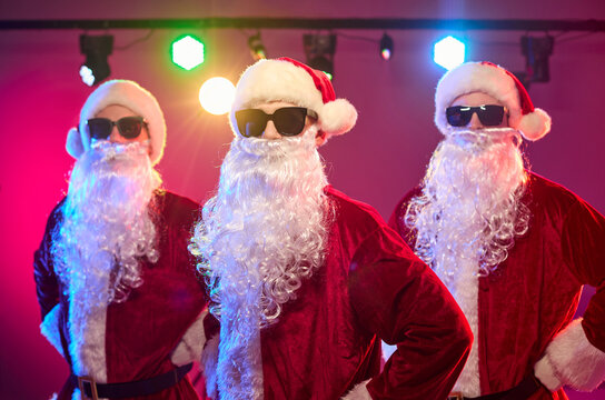 Santas dance at Christmas party in costumes. A trio of Santa dancers wears sunglasses and beards under bright vivid neon stage lights. Festive holiday party celebration and dance concept.