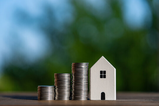 Property investment and financial growth concept with coin stacks and house model. Small white house model stands next to rising stacks of coins on wooden table with blurry green nature background