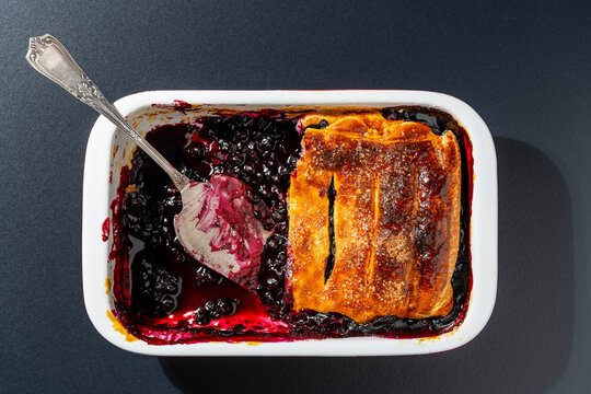 Homemade berry cobbler pie in baking dish with serving spatula overhead view, closeup. Delicious baked blueberry pie in white ceramic pan with a vintage metal spatula on dark background