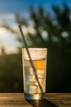 Refreshing cold drink with ice cubes and straw at sunset outdoor, closeup. Chilled glass of iced beverage with condensation droplets and drinking straw on wooden table over blurry sunset background