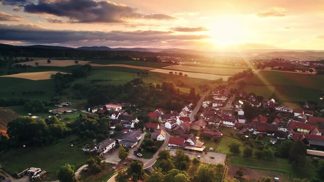 Aerial panning over a scenic village and rural landscape with hills, meadows, woods, the sun and illuminated house roofs at sunrise.