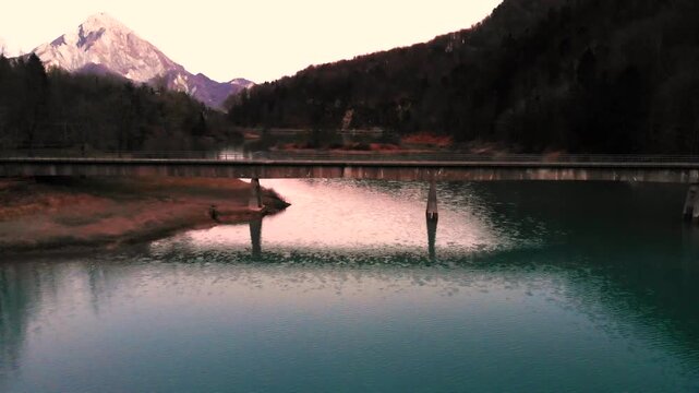 Horizontal 4K 24fps aerial drone video rising vertically over a road bridge to reveal the turquoise water of Lake Vergenis, with sunset reflections, a small pine islet, and winter mountains in Italy