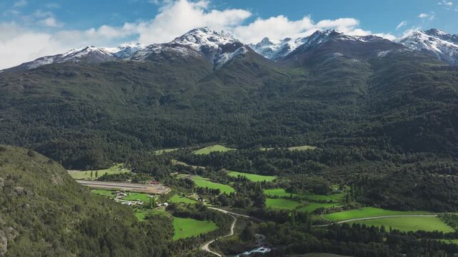 Aerial view of a remote town and airstrip nestled in a green valley, surrounded by dense forests and rugged snow-capped mountain ranges.
