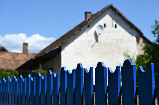 painted blue wood picket fence. diminishing perspective. decorative fence by the sidewalk. blurred white stucco finished old village type house beyond. sloped clay roof. cottage living concept