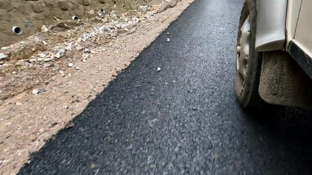 A low-angle, front wheel POV action shot of a 4x4 off-road vehicle driving along a newly paved asphalt highway in the rugged Himalayan terrain.