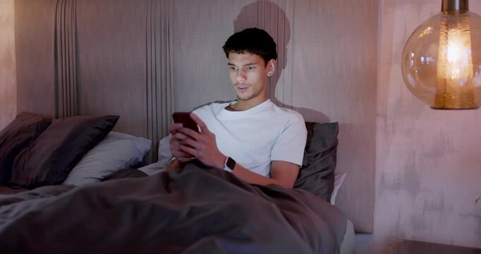 African American man holding smartphone, checking smartwatch, reading and smiling in bed under lamp