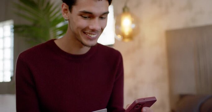 Adult male wearing maroon sweater checking card, holding phone, tapping, confirming payment on bed