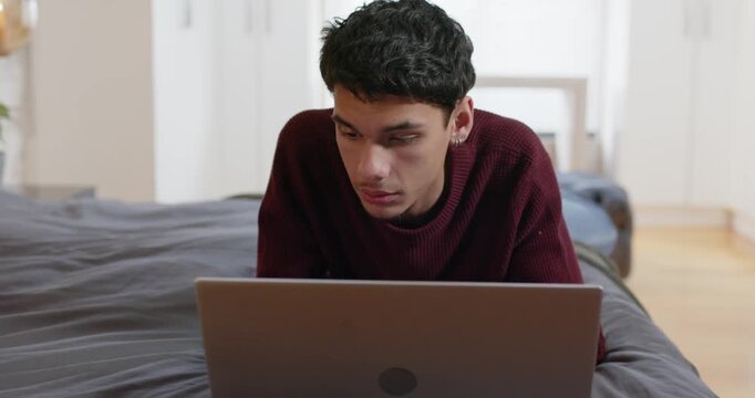 African American man propping on elbows, leaning and shifting gaze at laptop, studying in bedroom
