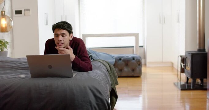 Adult male noticing laptop, propping on elbows on bed, reading, typing, pondering while working