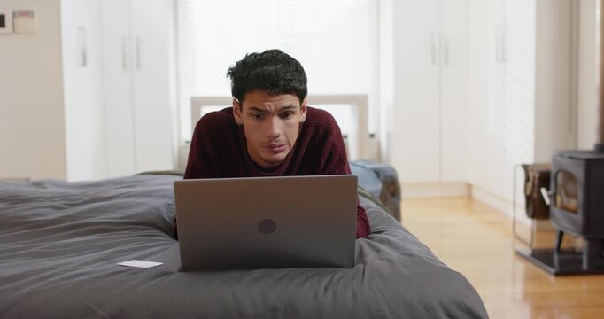African American man propping on elbows, lying on bed, studying on laptop, wearing burgundy sweater