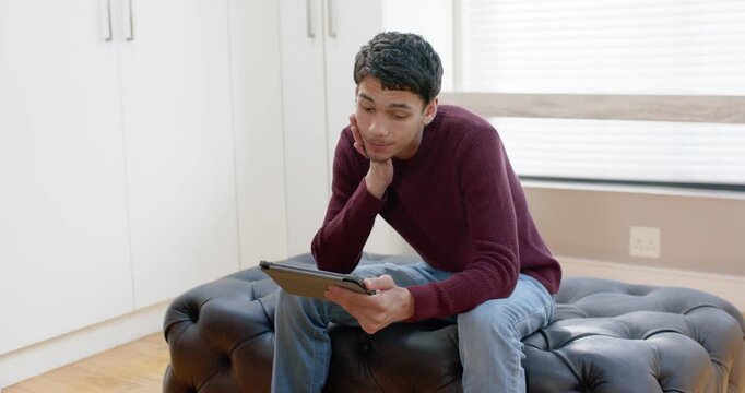 African American man in maroon sweater on ottoman at home glancing then tapping tablet reading
