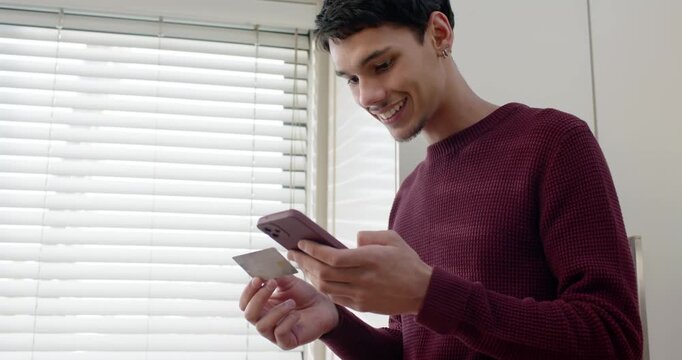 African American man in sweater holding phone, card, tapping, entering info at home near blinds