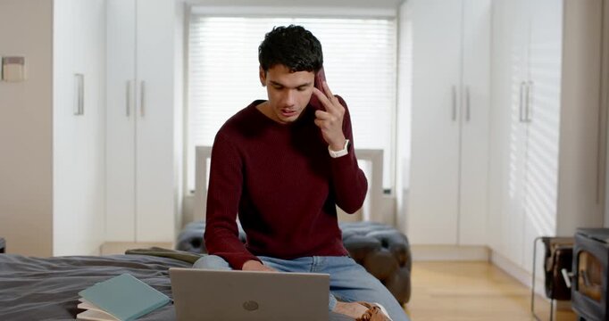 African American man in maroon sweater answering phone while typing on laptop in bedroom for work
